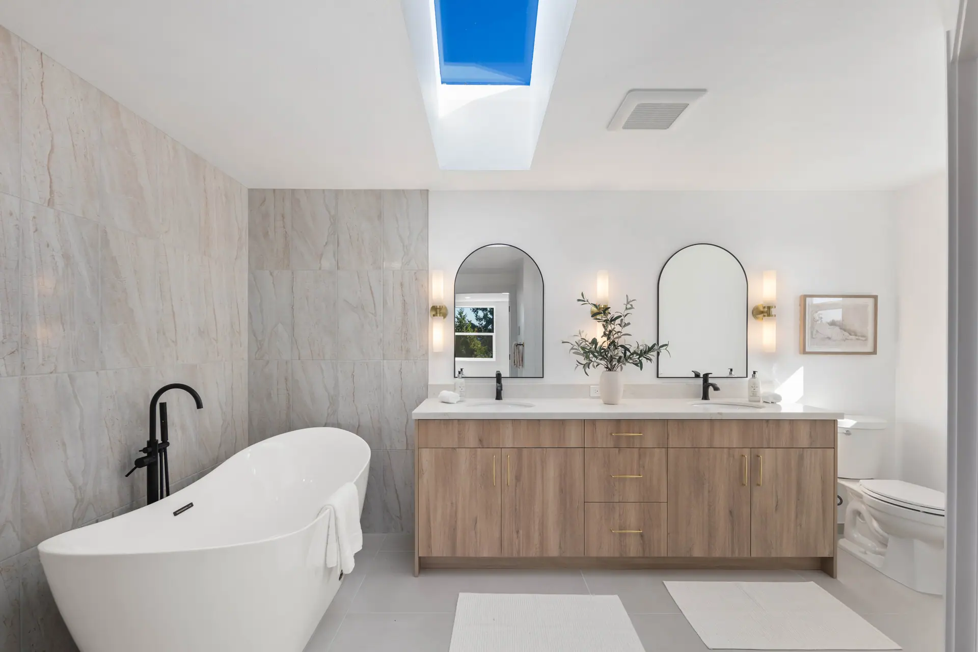 Bathroom with a freestanding tub, double vanity with light wood cabinets, arched mirrors and large-format stone-look tile
