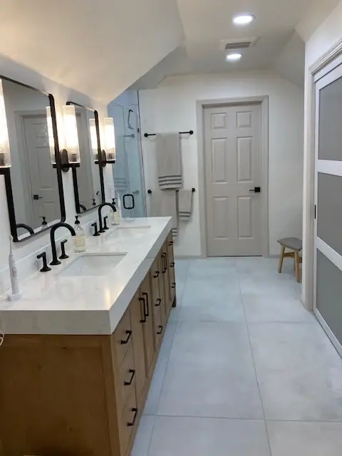 Remodeled primary bathroom with a light wood double vanity, black-framed mirrors, matte black faucets and large-format floor tile