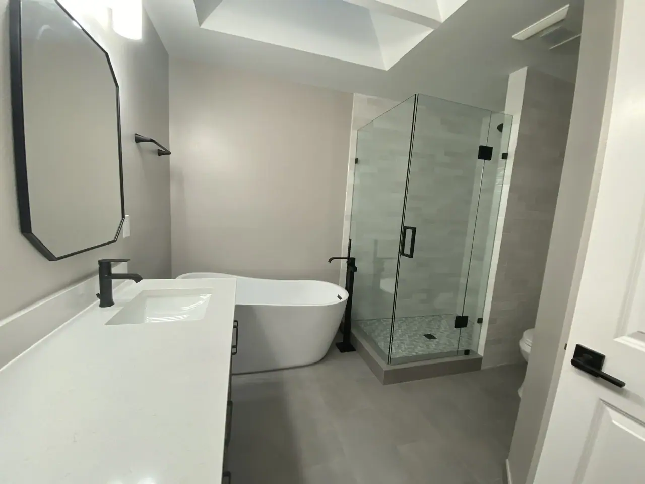 Bathroom under a skylight with a freestanding tub, a frameless glass corner shower, a white-top vanity and matte black fixtures
