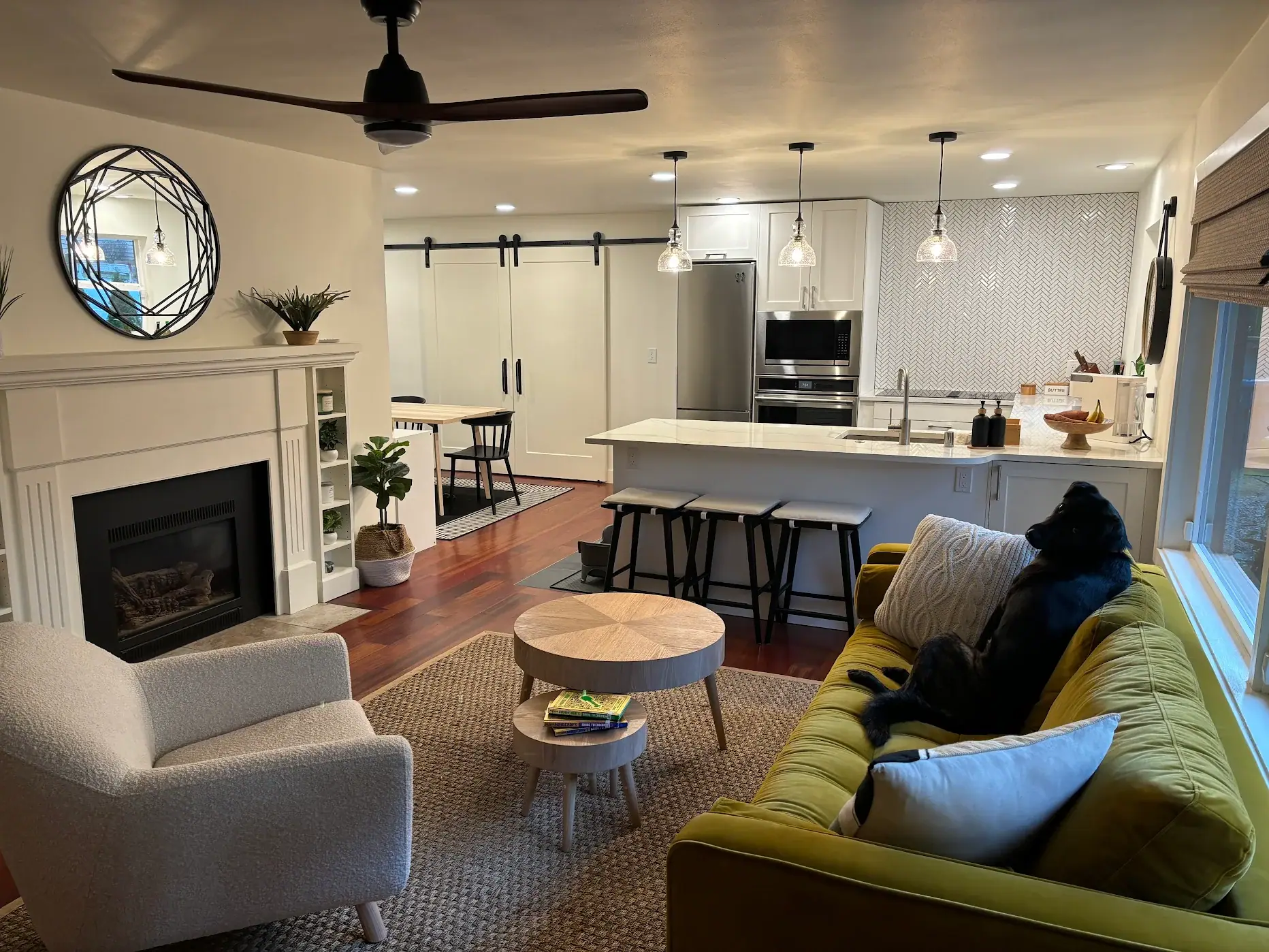 Living room with a fireplace open to a white kitchen with pendant lights and a herringbone tile backsplash