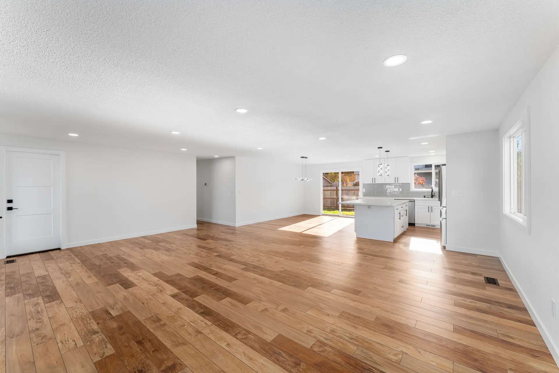 Open living area with wood-look flooring, recessed lighting and white walls leading to the kitchen
